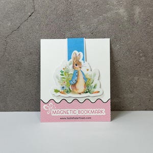 May include: A magnetic bookmark featuring a watercolor illustration of Peter Rabbit in a blue jacket, surrounded by flowers and carrots. The bookmark has a blue ribbon and a pink and white patterned base with the text "MAGNETIC BOOKMARK".