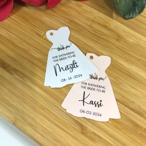 Puede incluir: Dos etiquetas de agradecimiento en forma de vestido, blancas y rosas, con el texto "gracias" y "POR DUCHAR A LA FUTURA NOVIA" y los nombres "Metzili" y "Kassi" con fechas.