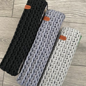 May include: Three crocheted storage baskets in black, grey, and light grey. Each basket has a brown leather tag with the word "studio" on it.