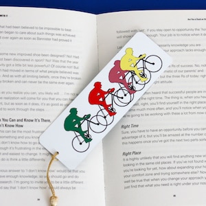 May include: A colourful bookmark featuring a design of four cyclists riding bicycles. The bookmark is made of paper and has a tassel. The text on the bookmark reads "Think You Can and Know It's There, But Don't Know How".