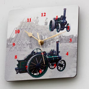 May include: Square wall clock with a vintage steam tractor design. The clock face has a grayscale background with two tractor illustrations and red numerals. The clock hands are gold-coloured.