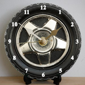 May include: A black and silver clock shaped like a car tyre with white numbers 1 through 12 around the rim. The clock hands are white and gold.