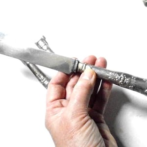 May include: A silver butter knife with an ornate handle. The knife is held in a hand.