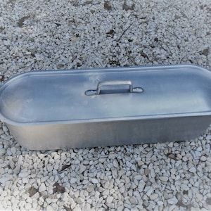 Puede incluir: Una fuente para asar de aluminio plateado con dos asas y una tapa. La fuente es de forma ovalada y tiene un solo asa en la tapa.