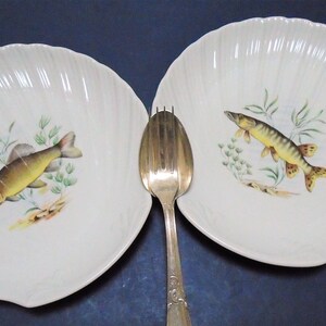 May include: Two white, scalloped-edge plates with a fish illustration and a silver fork. Each plate features a different fish design with green aquatic plants. The fork is positioned between the plates, creating a dining setting.