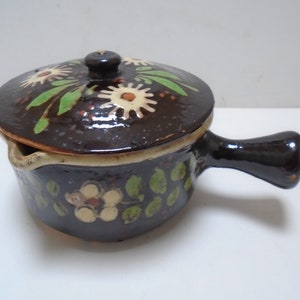 May include: A small brown ceramic pot with a lid, decorated with white and green floral designs. The pot has a handle and a spout.