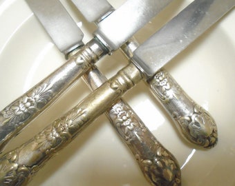 May include: Close-up of antique silverware knives with ornate floral designs on the handles. The blades are silver, and the handles have a tarnished, vintage appearance. The knives are arranged in a crisscross pattern.