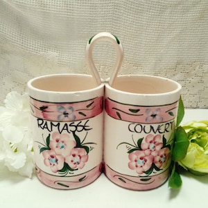 Puede incluir: Dos soportes para cubiertos de cerámica blanca con diseños florales rosas y verdes. Los soportes están conectados por un asa de metal. El soporte izquierdo dice "Ramasse" y el soporte derecho dice "Couvert".