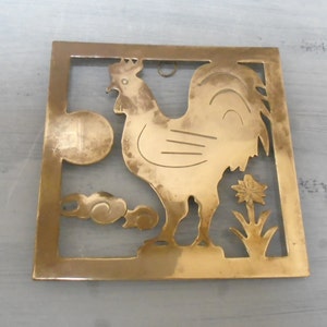 Peut inclure: Une découpe de coq en métal doré avec un motif décoratif. Le coq se tient sur une base avec une fleur et des nuages. La découpe est encadrée dans un carré.
