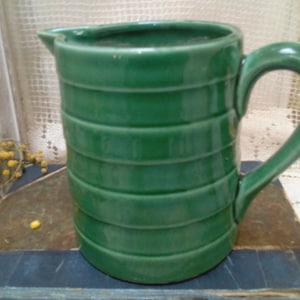 Puede incluir: Una jarra de cerámica verde con un diseño acanalado. La jarra tiene un asa y un pico.