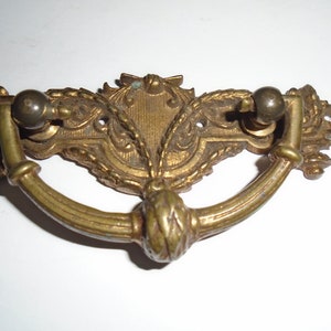 Può includere: Una maniglia per cassetto in ottone vintage con un design ornamentale. La maniglia ha un manico curvo con un pomo decorativo in basso e due pomelli più piccoli sui lati.