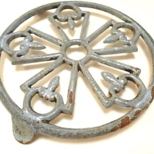 Może przedstawiać: Niebieski żeliwny podkładka pod gorące z dekoracyjnym wzorem. Podkładka ma okrągły kształt z uniesionym środkiem i sześcioma szprychami promieniującymi na zewnątrz. Szprychy są ozdobione wzorem kwiatowym. Podkładka ma trzy nogi.