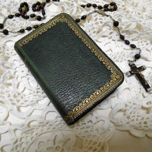 Puede incluir: Un libro encuadernado en cuero verde con detalles dorados y un rosario de madera marrón con un crucifijo de plata sobre un encaje blanco de ganchillo.