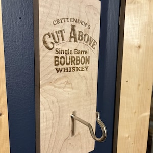 May include: A wooden sign with the text "Crittenden's Cut Above Single Barrel Bourbon Whiskey" in a brown font. A metal hook is attached to the bottom of the sign. The sign is mounted on a dark blue wall.