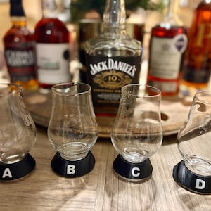 May include: Four clear, tulip-shaped whiskey tasting glasses with black bases labeled A, B, C, and D. The glasses are arranged on a wooden surface, with bottles of liquor in the background.