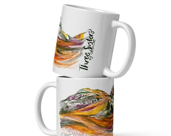 May include: Two white ceramic mugs stacked on top of each other. Each mug features a colorful landscape painting with the text "Three Sisters of Canmore". The artwork includes mountains, water, and vibrant colors.