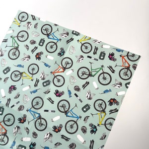 May include: A green, blue, and orange mountain bike pattern on a light green background. The pattern includes various cycling accessories, such as helmets, water bottles, and tools.