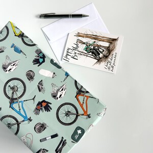 May include: A green and white gift wrap with a mountain bike design. The wrap features a variety of mountain biking accessories, including helmets, gloves, and tools. The text on the card reads "Happy Birthday! I heard it's all down hill from here."
