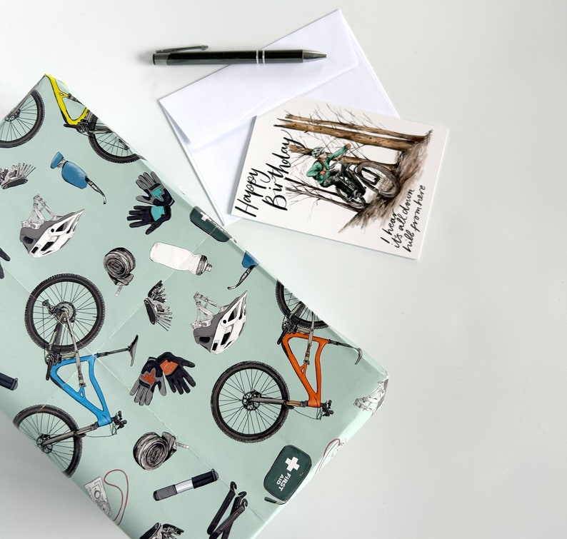 May include: A green and white gift wrap with a mountain bike design. The wrap features a variety of mountain biking accessories, including helmets, gloves, and tools. The text on the card reads "Happy Birthday! I heard it's all down hill from here."