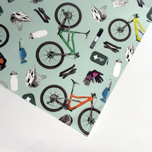 May include: A green and white patterned wrapping paper with a repeating design of mountain bikes, helmets, water bottles, and other cycling accessories. The text "FIRST AID" is printed on a small red and white box.