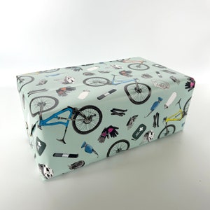 May include: A light blue gift wrap with a repeating pattern of mountain biking gear, including bicycles, helmets, gloves, water bottles, and tools.
