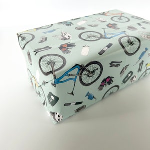 May include: A light blue gift wrap with a repeating pattern of mountain biking accessories, including bicycles, helmets, gloves, and water bottles.
