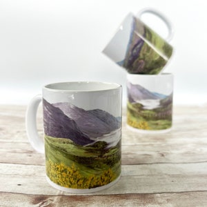 Può includere: Tre tazze in ceramica bianca con un design a paesaggio montano. Le tazze sono impilate l'una sull'altra. La tazza superiore presenta una catena montuosa viola con una valle verde. La tazza centrale presenta un lago blu con una valle verde. La tazza inferiore presenta un campo di fiori gialli con una valle verde.