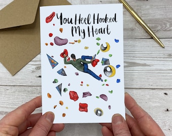 May include: A white card with a drawing of a person rock climbing on a wall with colorful holds. The text "You Heel Hooked My Heart" is written in black cursive.