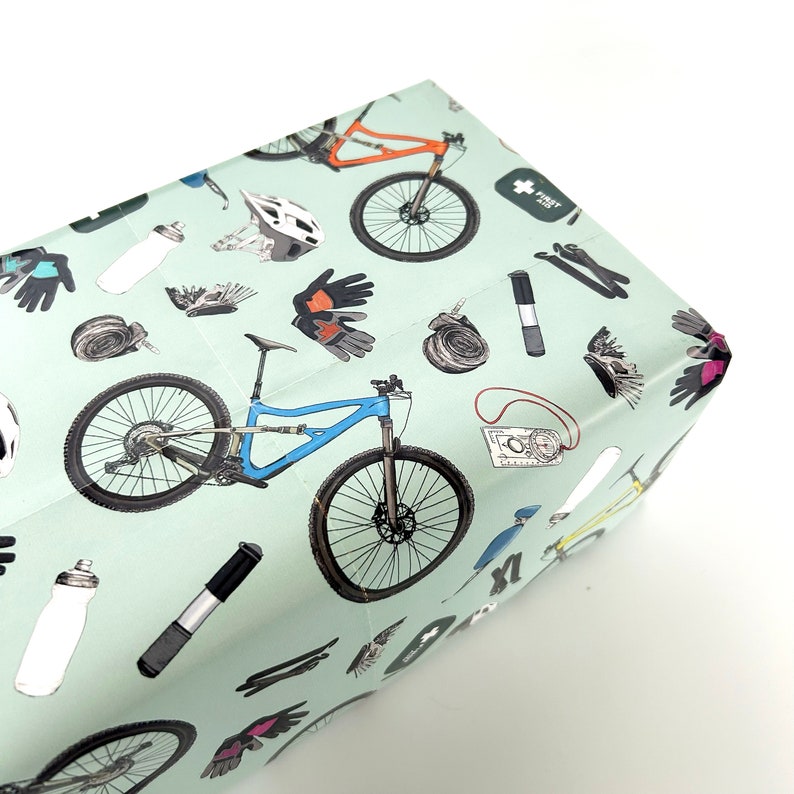 May include: A light green wrapping paper with a repeating pattern of mountain bikes, helmets, gloves, and other cycling accessories. The text "FIRST AID" is printed on a small red cross in the upper right corner.
