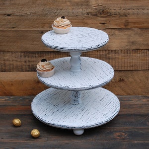 May include: White three-tiered cake stand with distressed paint finish. The stand has a round base and three circular tiers. The stand is made of wood and has a rustic look.