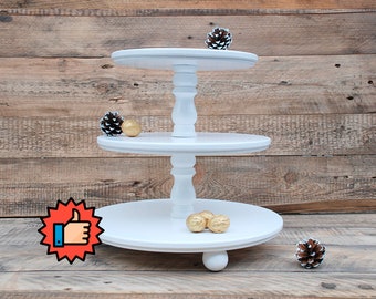 May include: White three-tiered cake stand with round platforms and turned wooden legs. The stand is decorated with pine cones and gold ornaments.