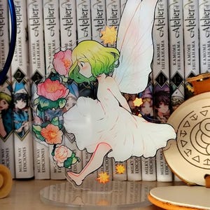 May include: A clear acrylic figure of a fairy with green hair, white dress, and delicate wings, surrounded by pink and orange flowers. The figure is on a clear stand, with a backdrop of books.