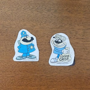 May include: Two vintage Cookie Crisp cereal box character stickers. One sticker features a police officer with a blue uniform and a star on his chest. The other sticker features a thief with a bag of cookies labeled "Cookie Crisp".