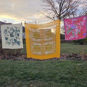 May include: Three colorful scarves hang on a clothesline in a backyard. The scarves feature various patterns: a floral design, a geometric pattern, and a botanical print. The scene is set against a backdrop of a house and a cloudy sky.