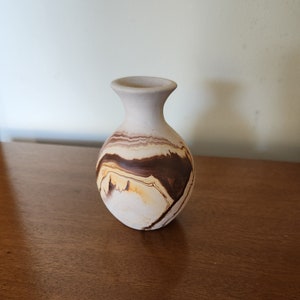 May include: A small, white ceramic vase with brown and yellow marbling. The vase has a rounded body and a narrow neck.
