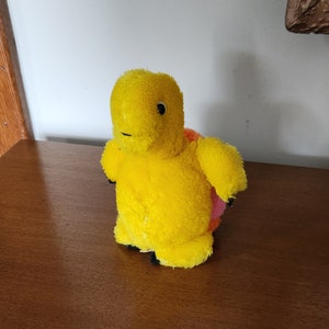 May include: A yellow plush turtle toy with a pink shell. The turtle has black eyes and a small black nose.