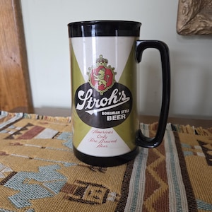 May include: A vintage beer stein with a black handle and rim. The stein features the Stroh's logo in a shield shape with a lion and crown. The text "Bohemian Style Beer" and "America's Only Fire Brewed Beer" are also visible.