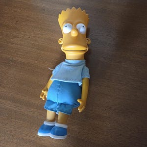 May include: A Bart Simpson doll with yellow skin, spiky yellow hair, and large white eyes. He wears a light blue t-shirt, blue shorts, and blue shoes with white trim. The doll is a collectible item.