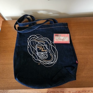 May include: A dark blue denim tote bag with white graphic text that reads "here and there". A small rectangular card with a landscape design is next to the bag. The bag has two denim handles.