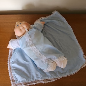 May include: A Cabbage Patch Kid doll wearing a light blue dress with white lace trim. The doll is lying on a light blue blanket with white lace trim.