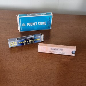May include: Three vintage sharpening stones in their original boxes. The boxes are labeled "Carborundum Pocket Stone", "Pocket & Desk Stone", and "CarboMagneto". The boxes are blue, silver, and tan.