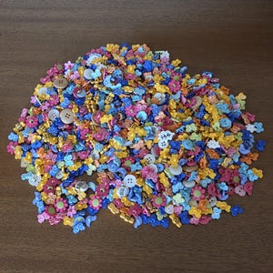 May include: A colorful assortment of craft buttons in various shapes and sizes, including flowers, butterflies, and round buttons. The buttons are in shades of pink, blue, yellow, and white, scattered on a brown surface.