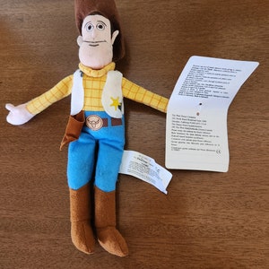 Op de afbeelding: Een pluche speelgoed van Woody uit de Toy Story-franchise. Woody draagt zijn kenmerkende bruine cowboyhoed, een geel geruit overhemd, blauwe jeans en bruine laarzen. Hij heeft een sheriffster-badge op zijn shirt en een bruine riem met een stierenkopgesp. Het speelgoed zit op een houten oppervlak.