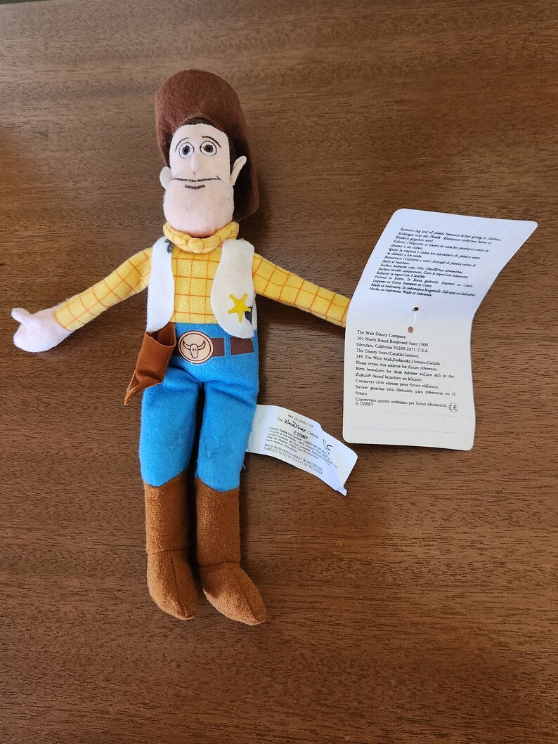 Op de afbeelding: Een pluche speelgoed van Woody uit de Toy Story-franchise. Woody draagt zijn kenmerkende bruine cowboyhoed, een geel geruit overhemd, blauwe jeans en bruine laarzen. Hij heeft een sheriffster-badge op zijn shirt en een bruine riem met een stierenkopgesp. Het speelgoed zit op een houten oppervlak.