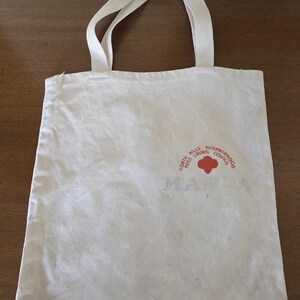 May include: Off-white canvas tote bag with two handles. The bag features a red and orange circular logo with the text "North Hills Laurel Neighborhood Council" and the word "MANDA" printed below the logo.