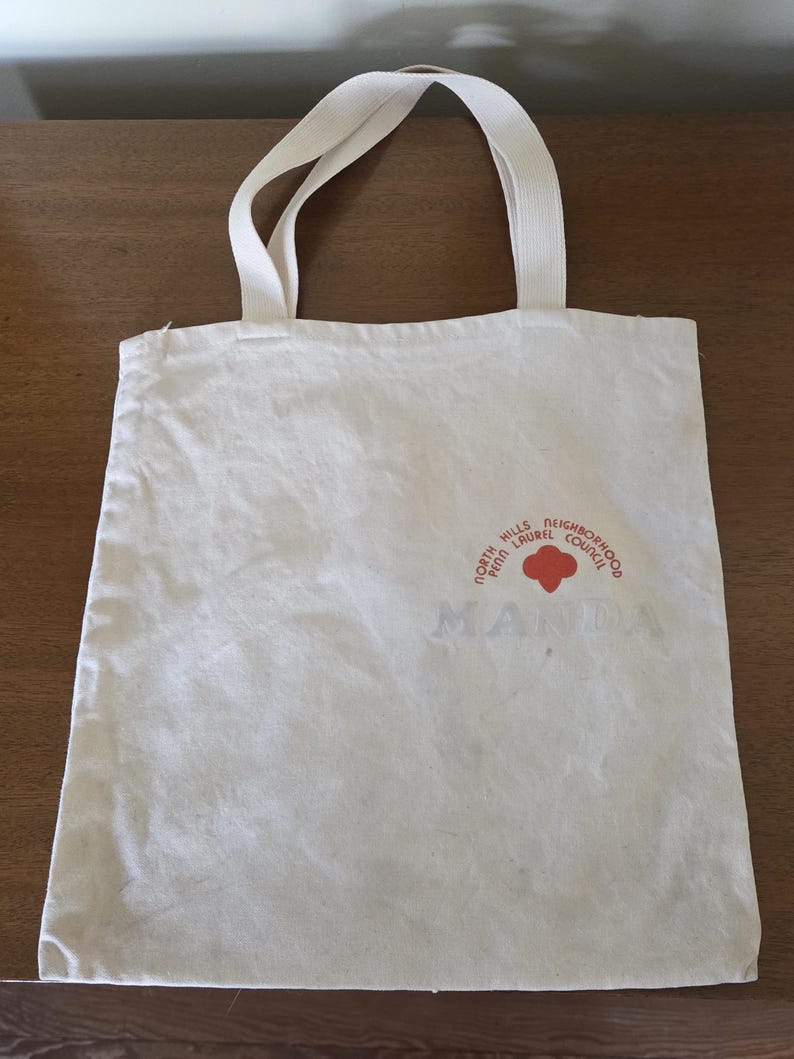 May include: Off-white canvas tote bag with two handles. The bag features a red and orange circular logo with the text "North Hills Laurel Neighborhood Council" and the word "MANDA" printed below the logo.