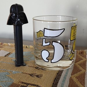 May include: A black Darth Vader PEZ dispenser stands next to a clear glass tumbler. The tumbler features a large white number "5" and cartoon graphics with the text "If you is n... 6,7... then you..."