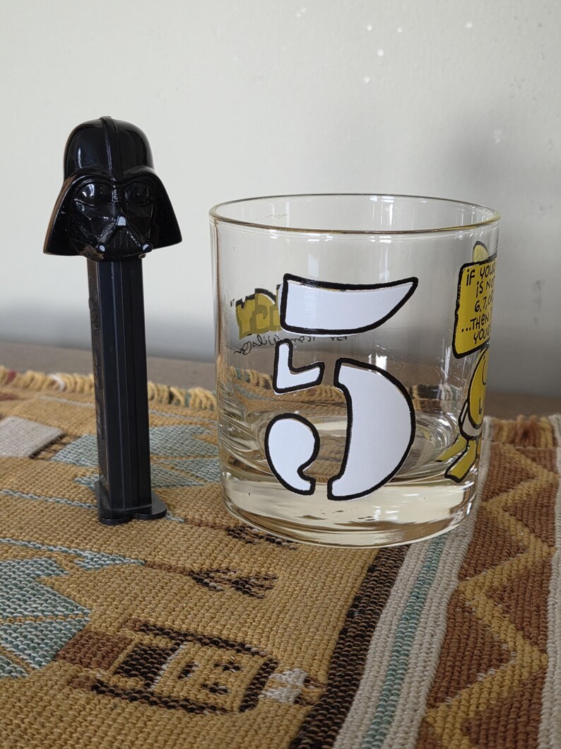 May include: A black Darth Vader PEZ dispenser stands next to a clear glass tumbler. The tumbler features a large white number "5" and cartoon graphics with the text "If you is n... 6,7... then you..."