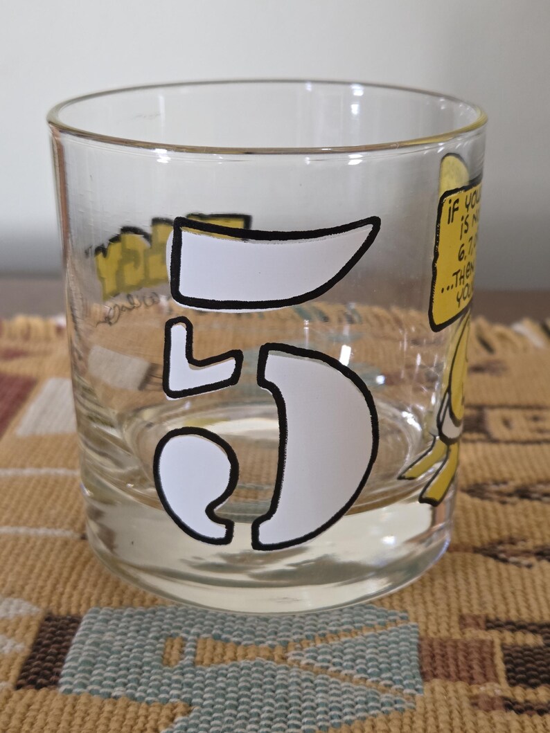 May include: A clear glass tumbler with a white outlined number "5" and yellow cartoon graphics. The glass has text that reads "If you is not 6.7...then you". The glass is on a patterned surface.