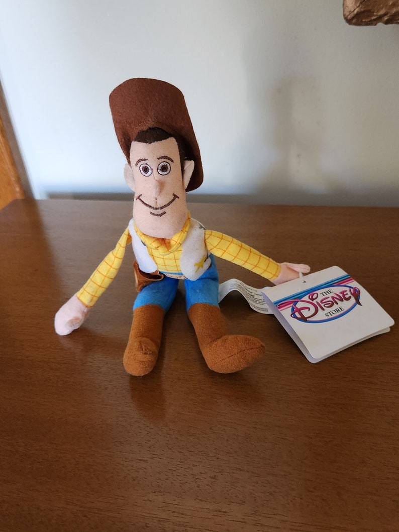 Op de afbeelding: Een pluche speelgoed van Woody uit de Toy Story-franchise, met zijn kenmerkende bruine hoed, gele shirt en blauwe vest. Het speelgoed zit met gekruiste benen en heeft een vriendelijke uitdrukking. Het speelgoed heeft een label met de tekst "The Disney Store".